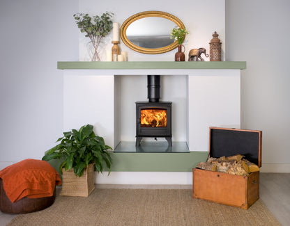 A modern wood-burning stove insert set into a white wall niche with sage green mantel and base shelves. The scene is styled as a cozy living room with a lit fire, a gold-framed mirror, decorative plants, a leather trunk, and an orange pouf on a woven rug.