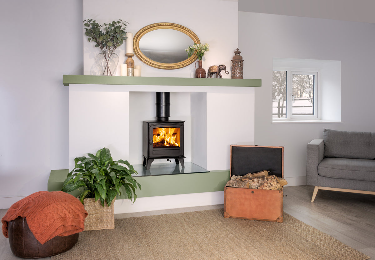 A modern wood-burning stove installed in a white and sage green fireplace niche, set in a styled living room with a gray sofa, a potted plant, and a rustic orange trunk.