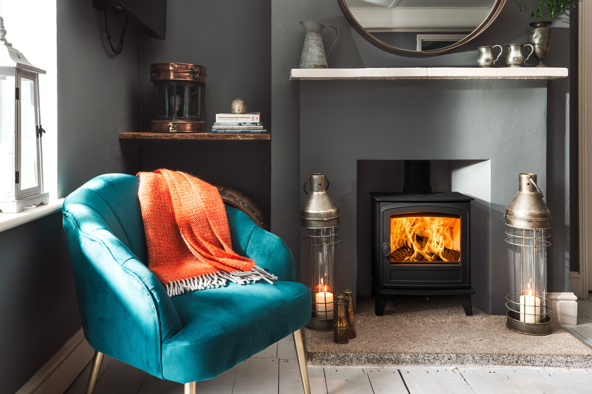 A cozy living room corner featuring a teal velvet armchair with an orange throw blanket, set beside a black wood-burning stove with a glowing fire. The scene includes a fireplace mantel with decorative items, a round mirror, and two brass lanterns on a tiled floor.