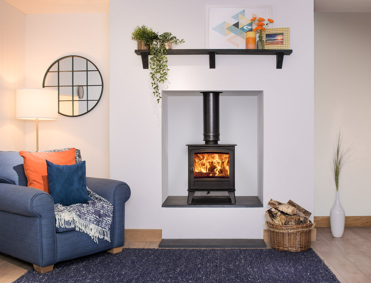 A black wood-burning stove with a visible fire, set into a white wall fireplace niche in a styled living room with a blue armchair, orange and blue pillows, a shelf with plants, and a basket of firewood on the floor.