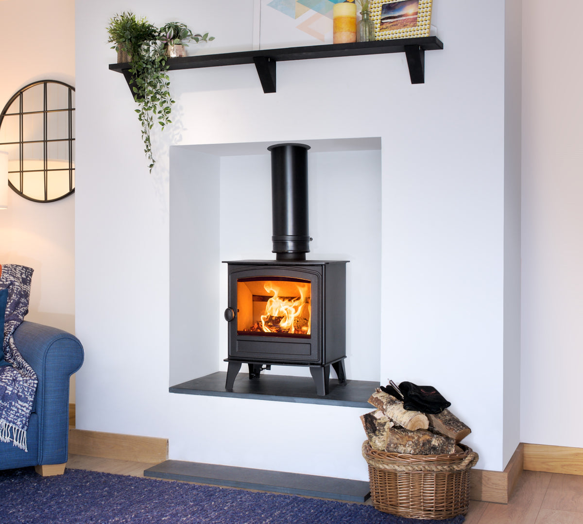 A black wood-burning stove with a visible fire, set into a white wall niche in a styled living room. A black shelf with plants and decor sits above, and a wicker basket of logs is on the floor below.