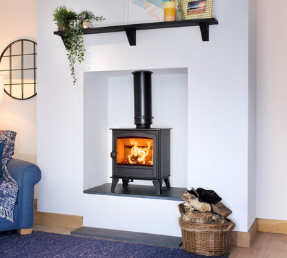 A black wood-burning stove with a visible fire, set into a white wall niche in a styled living room. A black shelf with plants and decor sits above, and a wicker basket of logs is on the floor below.