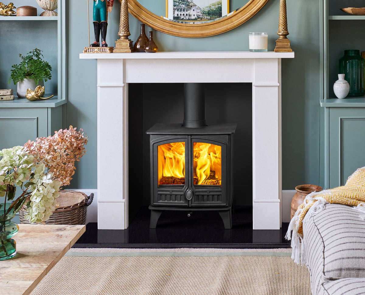 A black wood-burning stove with glowing orange flames set within a white fireplace mantel in a styled room with blue-green walls and home decor.