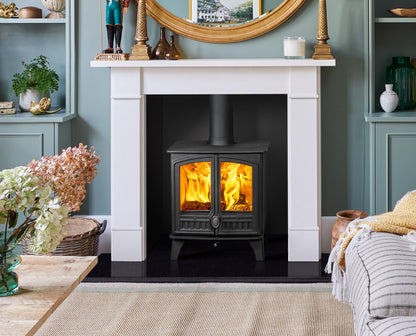 A black wood-burning stove with glowing orange flames set within a white fireplace mantel in a styled room with blue-green walls and home decor.