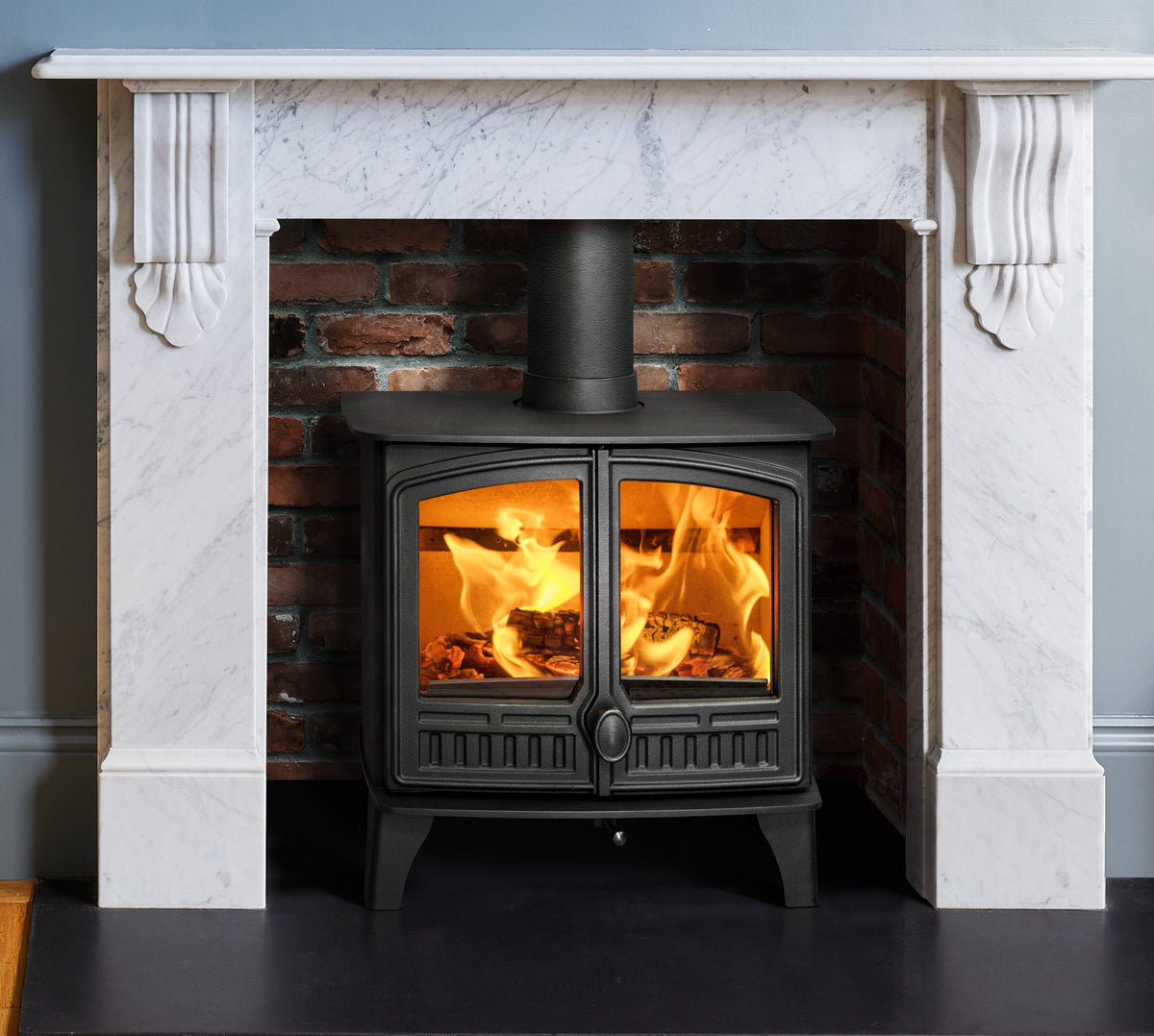A black wood-burning stove with glowing orange flames, set within a brick fireplace surround framed by a white marble mantel.