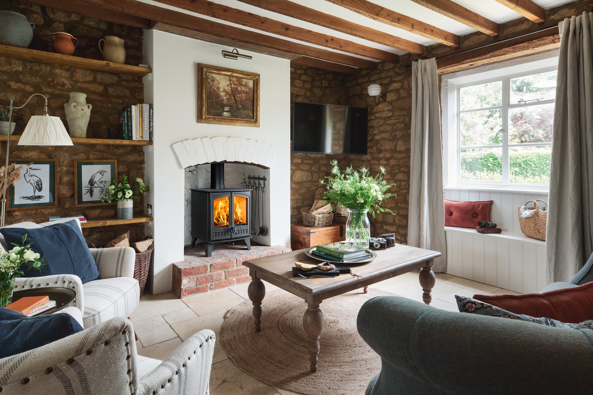 A cozy, rustic living room featuring a black wood-burning stove with a glowing fire, set against a white wall and brick fireplace. The room has exposed wooden ceiling beams, stone brick walls, a wooden coffee table, and comfortable armchairs.
