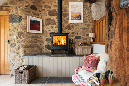A black wood-burning stove with a visible fire set into a stone wall in a rustic living room. A patterned armchair with a white faux-fur throw and pillows sits nearby, along with wicker baskets, framed wall art, and a large natural wood post.