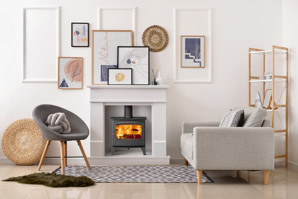 A modern living room scene featuring a black wood-burning stove with a glowing fire inside a white fireplace mantel. The room is styled with a gray accent chair, a light beige sofa, a patterned rug, a wooden shelving unit, and framed wall art.