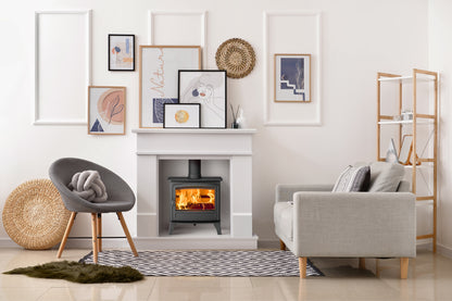 A modern living room scene featuring a black wood-burning stove with a glowing fire inside a white fireplace mantel. The room is styled with a gray accent chair, a light beige sofa, a patterned rug, a wooden shelving unit, and framed wall art.