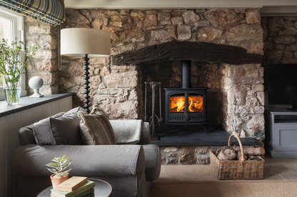 A black wood-burning stove with glowing orange flames, set within a stone fireplace surround with a solid wooden beam above in a cosy lounge setting.