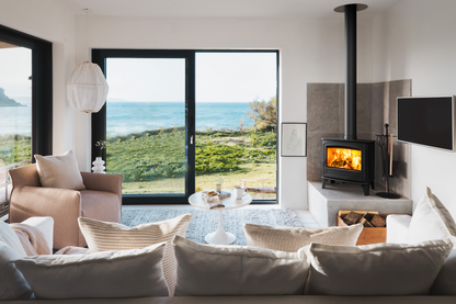A modern living room with a black wood-burning stove with a visible fire, set in a stone hearth. The room features a large window looking out onto a green hillside and the blue ocean, with a beige sofa and armchair in the foreground.