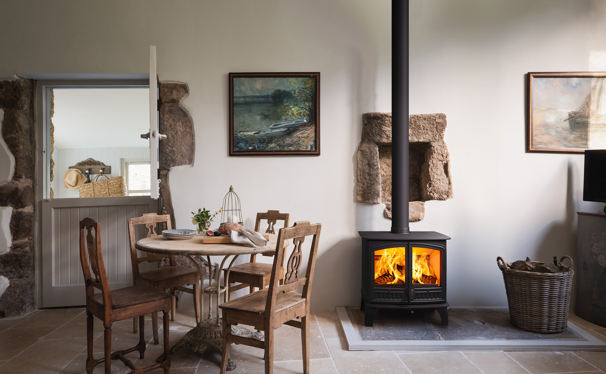 A rustic interior scene with a black wood-burning stove with visible flames, a round wooden dining table and chairs, stone walls, framed landscape paintings, and a basket on the floor.