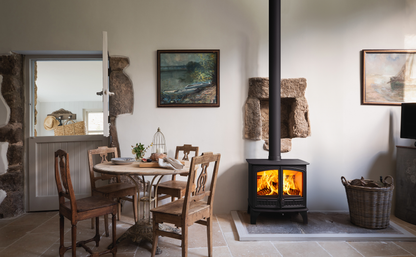A rustic interior scene with a black wood-burning stove with visible flames, a round wooden dining table and chairs, stone walls, framed landscape paintings, and a basket on the floor.