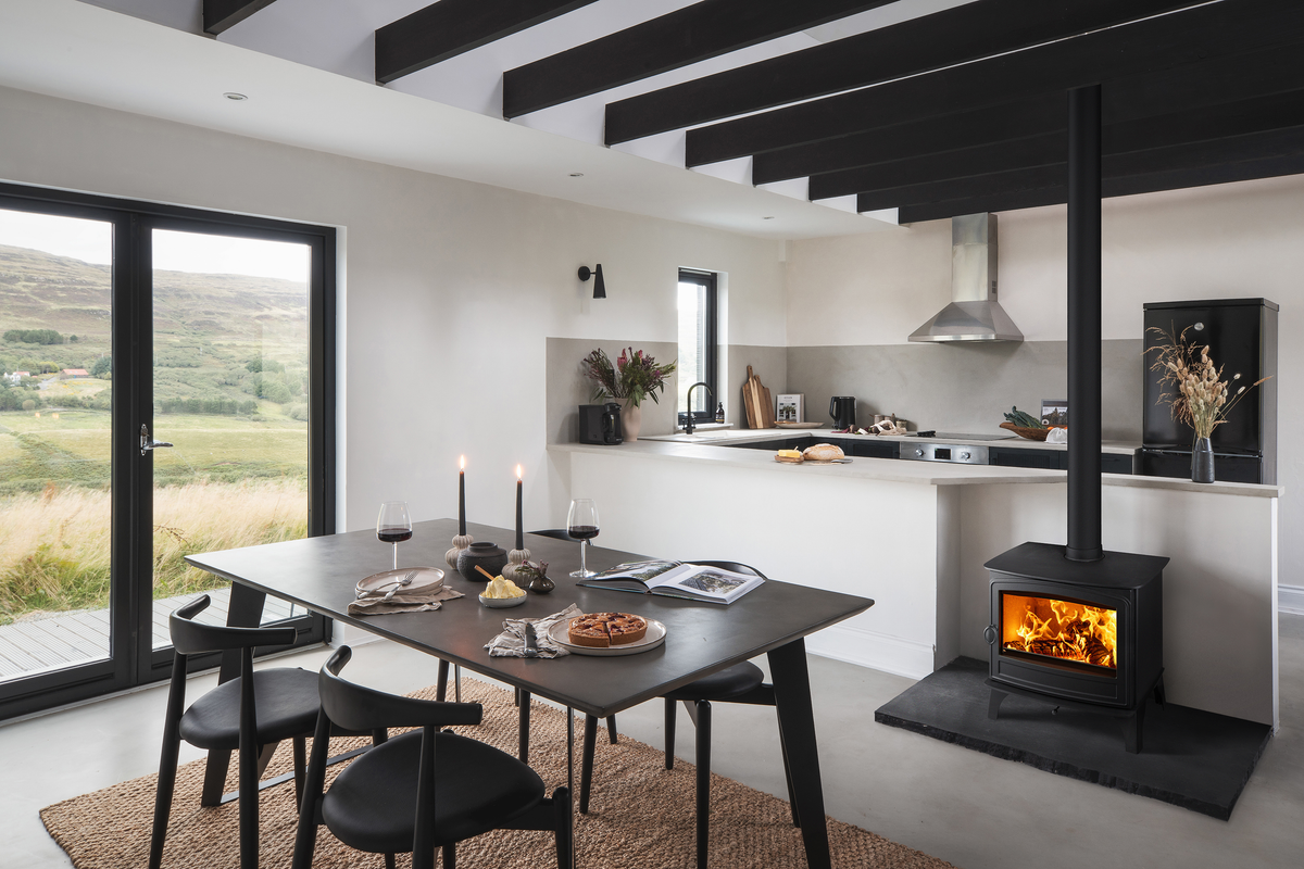 A modern interior room with a black dining table and chairs, a black wood-burning stove with visible flames, a kitchen in the background, and large windows showing a green landscape outside.
