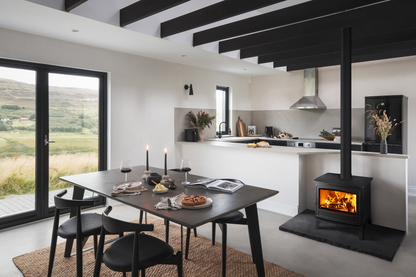 A modern interior room with a black dining table and chairs, a black wood-burning stove with visible flames, a kitchen in the background, and large windows showing a green landscape outside.