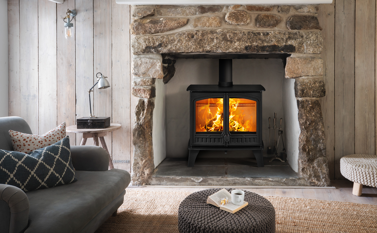 A black wood-burning stove with a glowing fire inside, set within a rustic stone fireplace in a cozy living room with a gray sofa and woven rug.