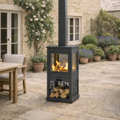 Outdoor patio heater with firewood storage on a stone patio, surrounded by potted plants and a building.