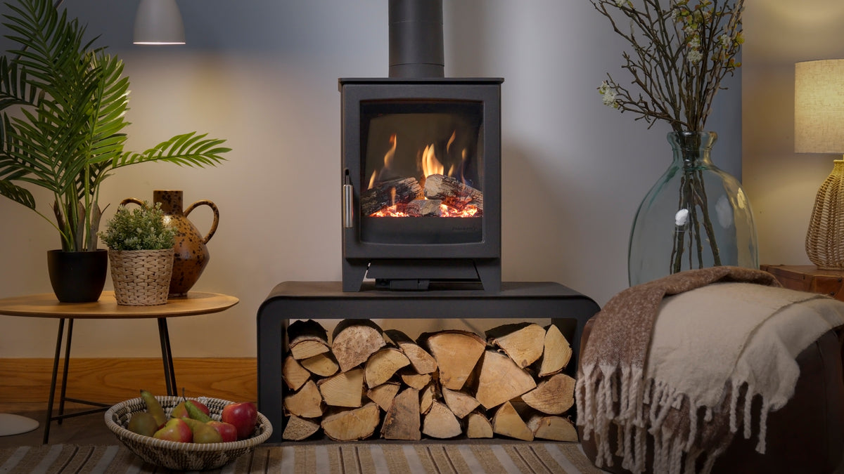 A black gas stove with a glass front door showing a glowing fire, set on a matching firebox shelf filled with stacked firewood. The scene is styled in a cozy living room with a side table, potted plants, a large glass vase with branches, and a throw blanket.