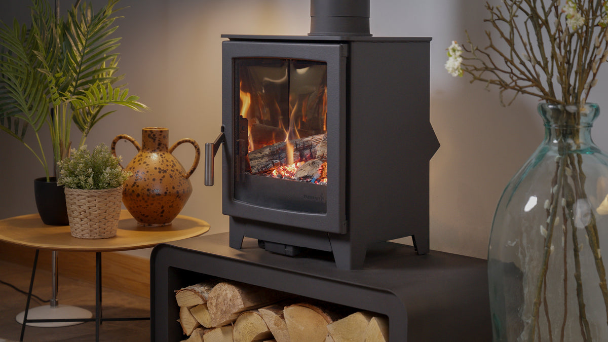 A black gas stove with a glass door showing a glowing fire, set in a cozy room next to a side table with plants and a ceramic pot.