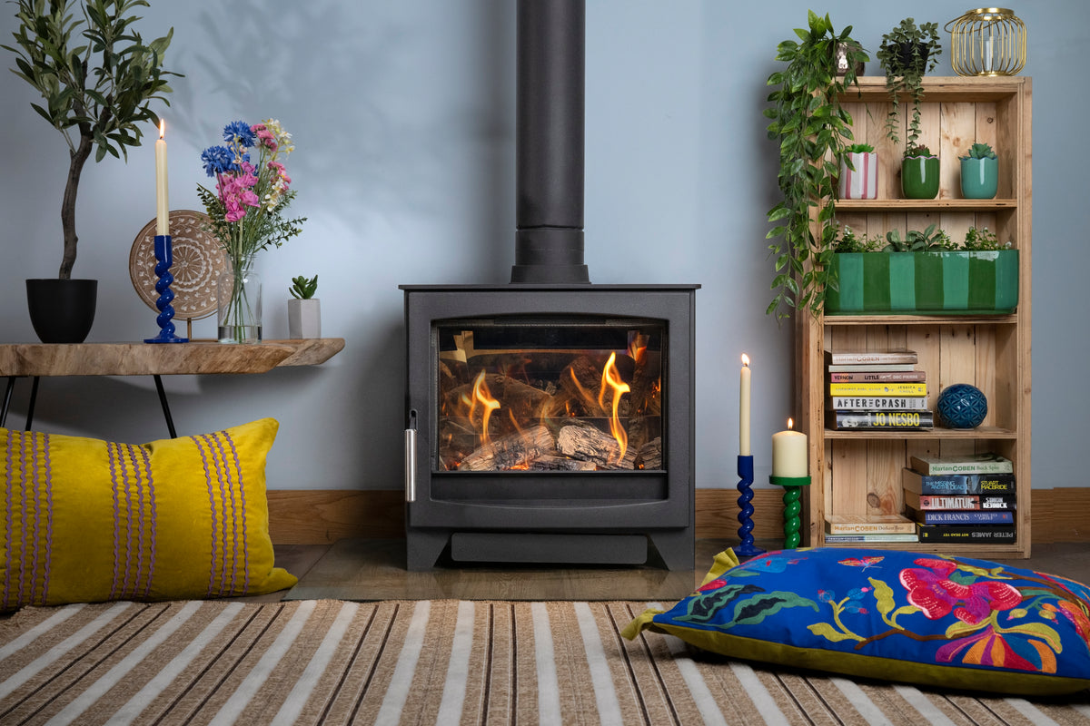 A black gas stove with a visible fire, set in a styled room with a blue wall, a striped rug, colorful pillows, a wooden shelf with plants and books, and a floating shelf with candles and flowers.