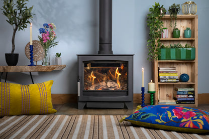 A black gas stove with a visible fire, set in a styled room with a blue wall, a striped rug, colorful pillows, a wooden shelf with plants and books, and a floating shelf with candles and flowers.