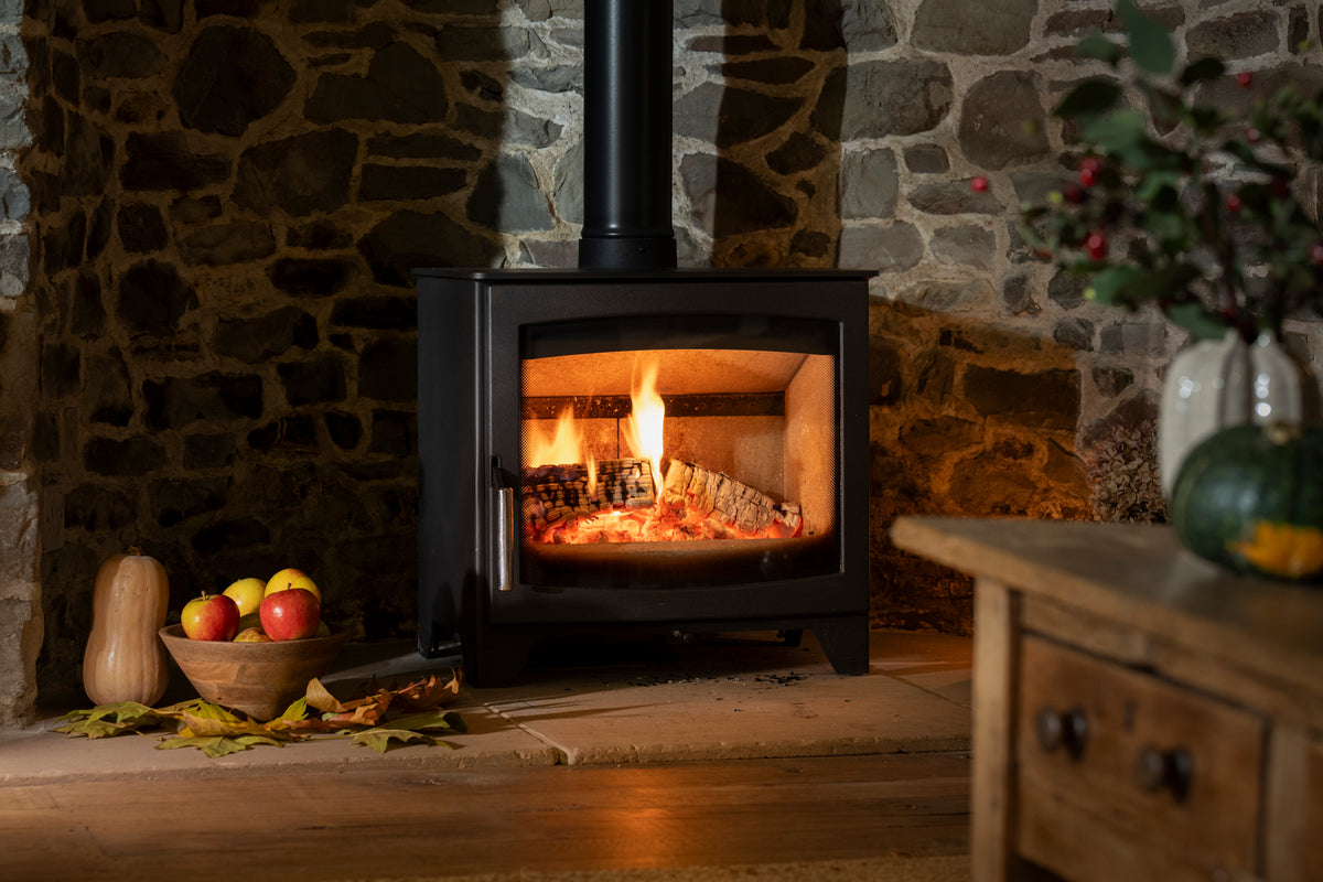 A black wood-burning stove with a visible fire inside, set against a rustic stone wall in a cozy room scene with a wooden table and bowl of fruit nearby.