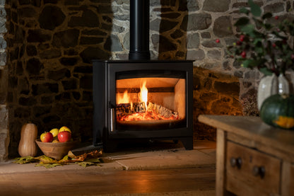 A black wood-burning stove with a visible fire inside, set against a rustic stone wall in a cozy room scene with a wooden table and bowl of fruit nearby.