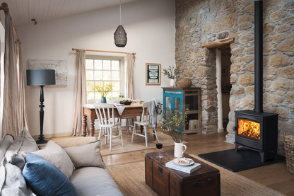 A cozy rustic living room featuring a black wood-burning stove with a glowing fire, a stone fireplace wall, a light gray sofa with blue pillows, and a wooden dining table in the background.