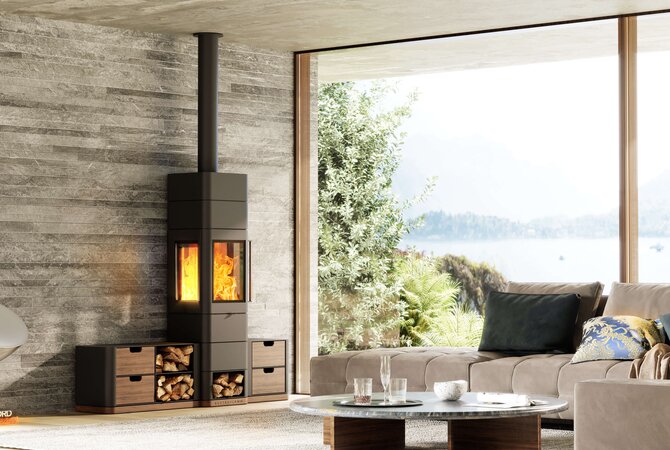 A modern black wood-burning stove with a visible fire, set against a textured gray stone wall in a contemporary living room with a large window overlooking water and trees.