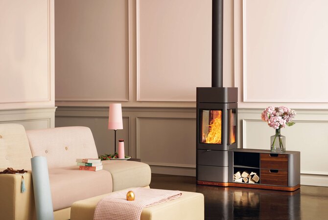 A modern wood-burning stove with a tall black chimney stack, set in a styled living room next to a cream sofa and a small wooden sideboard with flowers.