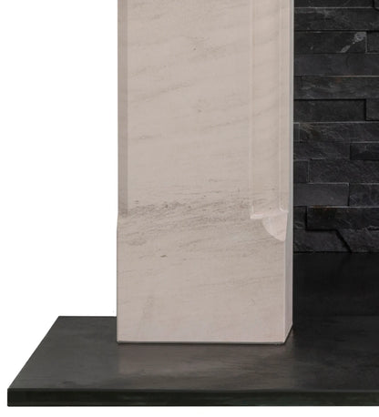A close-up of a rectangular stone fireplace in a light warm grey / beige tone, standing upright on a dark charcoal surface against a dark stacked-stone wall background.