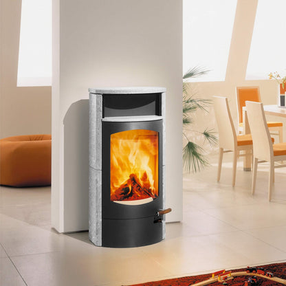 A modern wood-burning fireplace insert with a black front body, textured gray side panels, and a glowing orange fire visible through the glass door, set in a bright contemporary room.