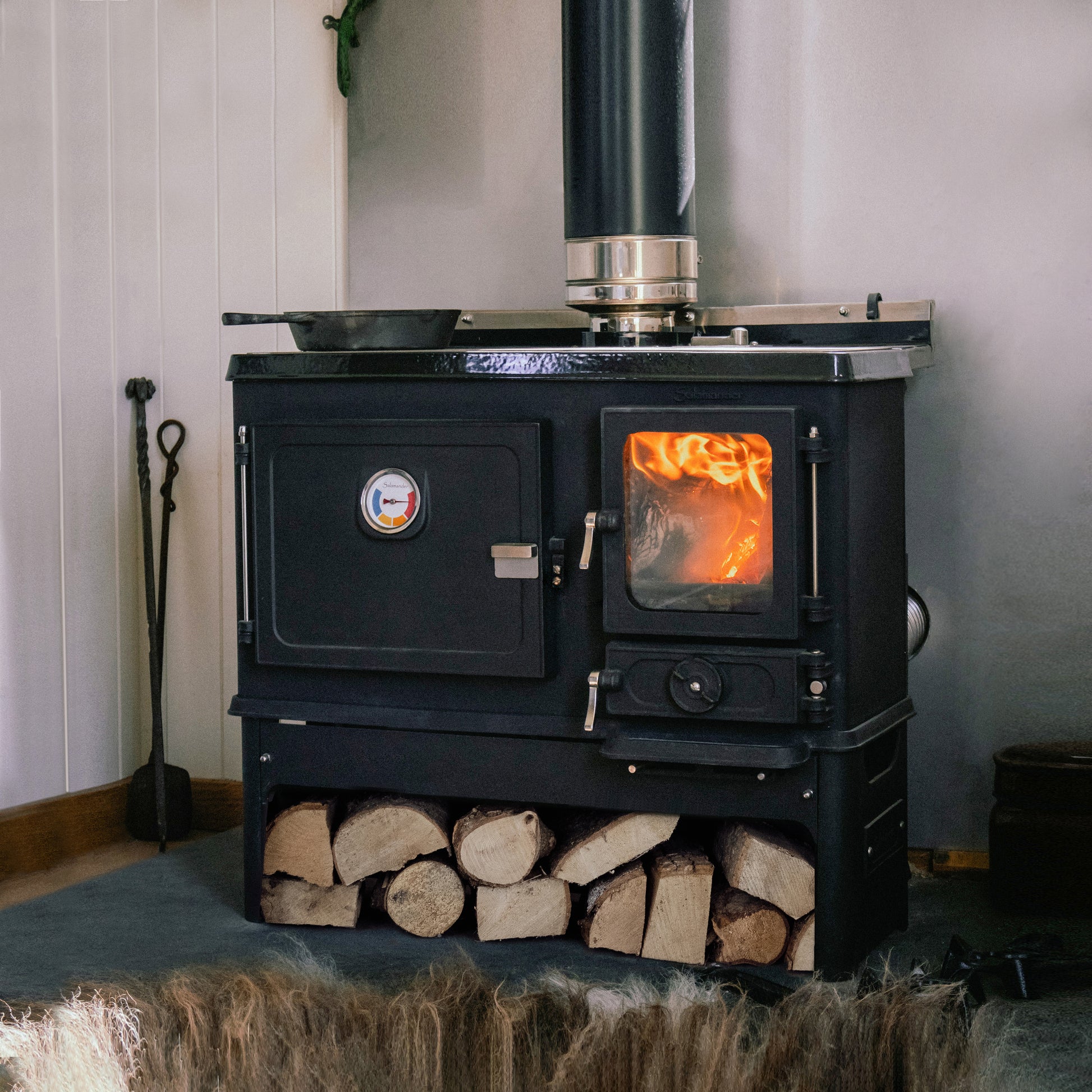 Salamander Little Range Cookstove Black wood stove with glowing interior and stacked logs in a room with white walls.