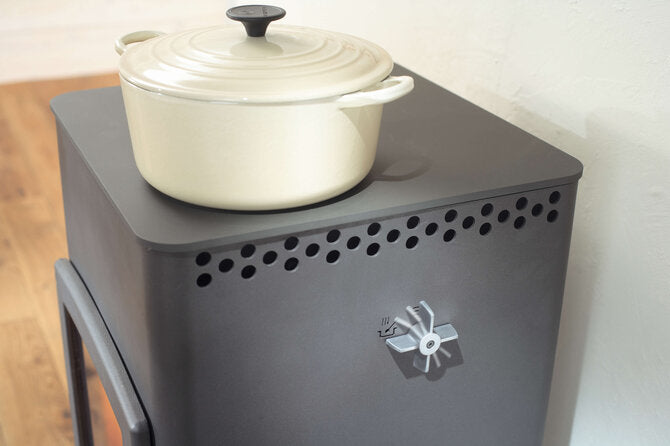 A dark gray metal wood-burning stove with a cream-colored enameled pot on top, set against a plain white wall and light wood floor.