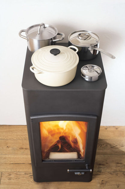 A dark gray wood-burning stove with a visible fire inside, topped with several cookware pots and a lid on a light wood floor against a plain white wall.