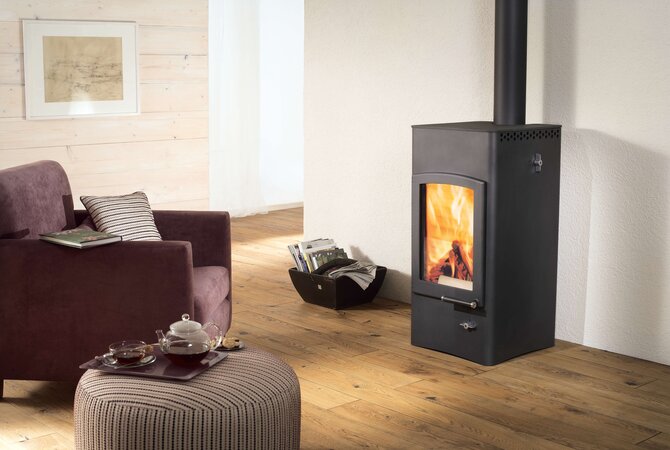 A black wood-burning stove with a visible fire, set in a cozy living room with a wooden floor, a purple armchair, and a striped ottoman.