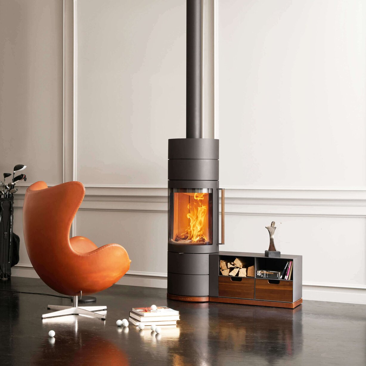 A modern dark gray wood-burning stove with a visible fire, set in a styled room next to an orange chair and a small wooden cabinet.