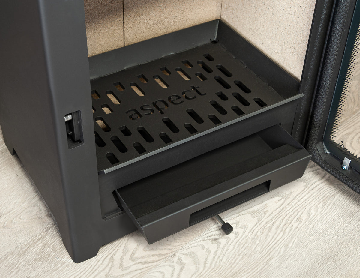 Close-up of a dark charcoal gray Aspect fireplace insert, showing the interior firebox with a perforated grate and a sliding drawer pulled open, set against a light wood floor and textured wall.