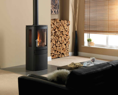 A black gas stove with a visible fire, set in a modern living room next to a window with blinds and a stack of firewood.