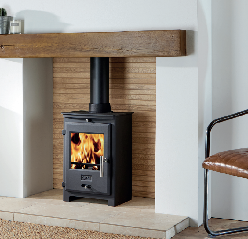 OER 5kW Multi Fuel Stove. Wood-burning stove in a modern room with wooden mantel and chair.