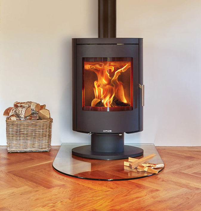 Opus Harmony PR Wood-burning stove showing flames, placed on a glass hearth with wooden floor with a basket of wood logs beside it.