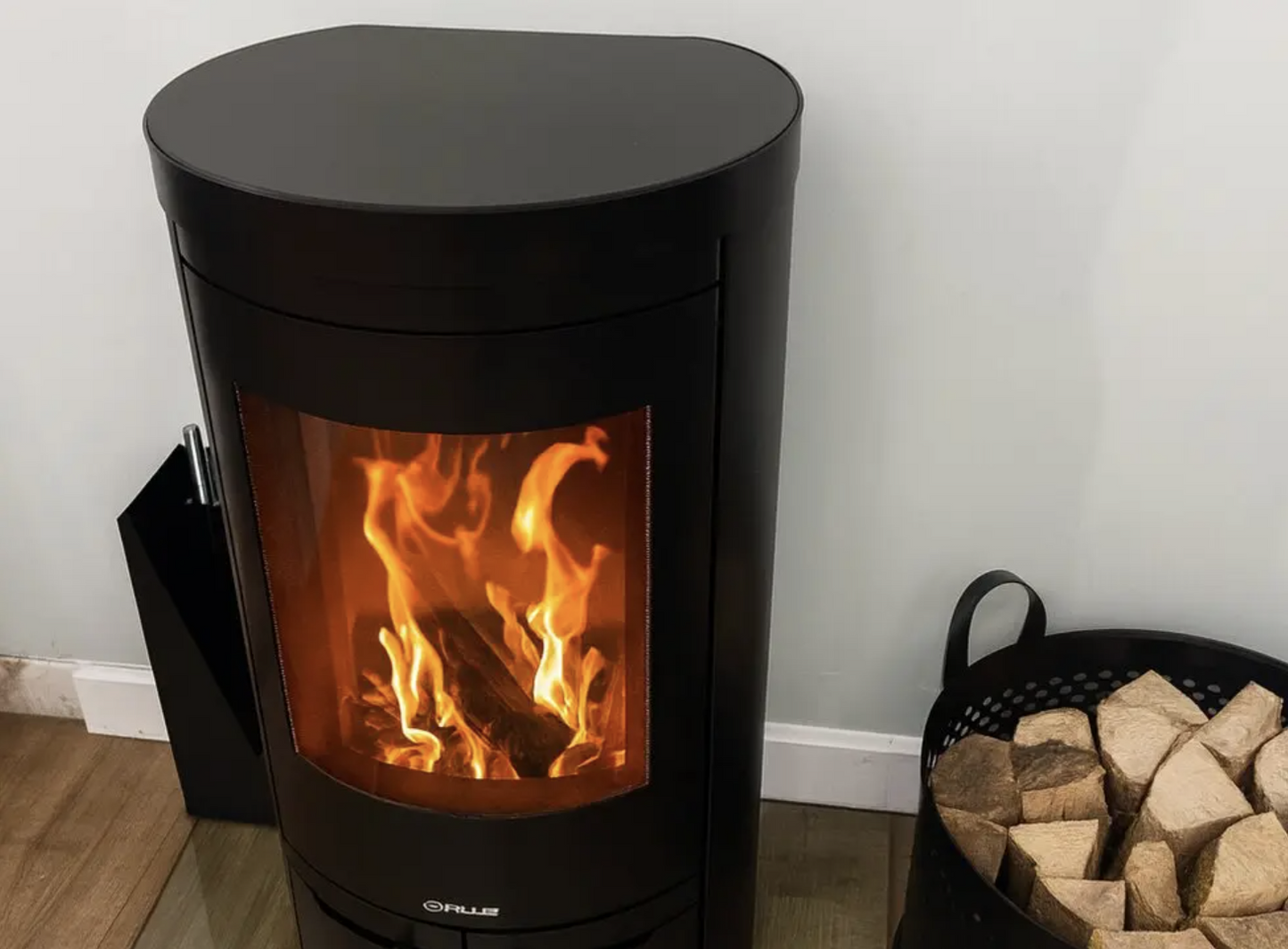 Opus Aria Black wood-burning stove with flames inside, next to a basket of wood on a wooden floor.