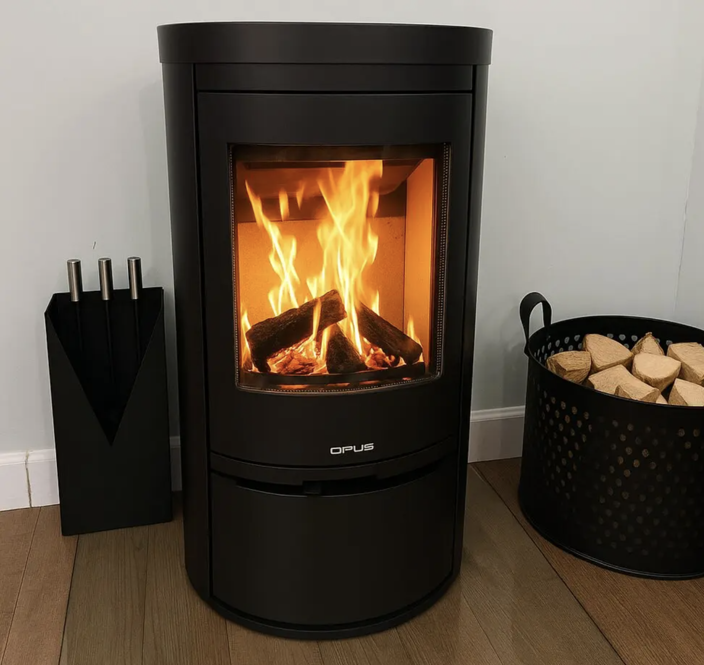 Opus Aria Black stove with flames inside, next to a basket of wood and a knife block on a wooden floor