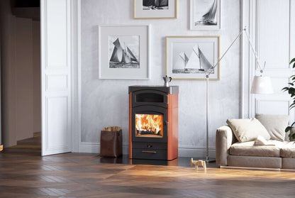 A modern wood-burning stove with a visible fire, set in a styled living room with a gray wall, framed sailboat prints, a beige sofa, and a floor lamp.
