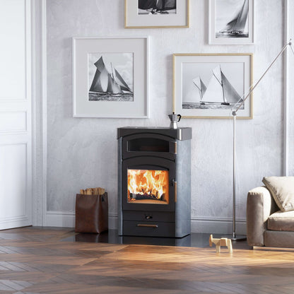 A black wood-burning stove with an open fire, set against a light gray wall in a styled room with framed sailboat prints, a floor lamp, and a sofa.