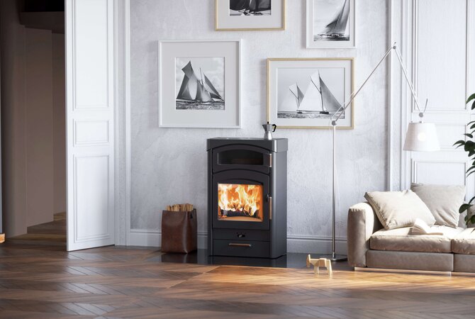 A black wood-burning stove with a glowing fire, set in a modern living room with a gray wall, framed sailboat prints, a beige sofa, and a floor lamp.