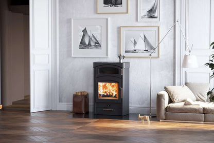 A black wood-burning stove with a glowing fire, set in a modern living room with a gray wall, framed sailboat prints, a beige sofa, and a floor lamp.