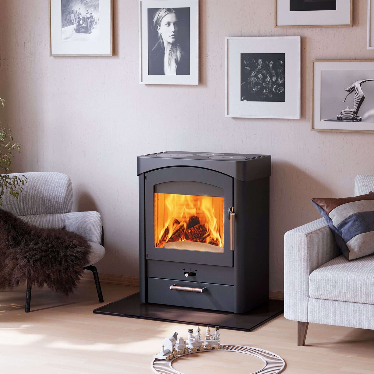 A black wood-burning stove with a glowing orange fire inside, set in a modern living room next to two gray armchairs against a beige wall with framed black-and-white photos.