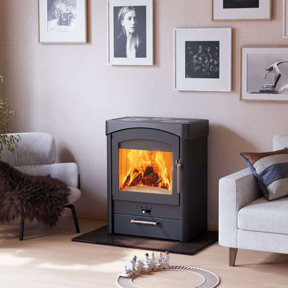 A black wood-burning stove with a glowing orange fire inside, set in a modern living room next to two gray armchairs against a beige wall with framed black-and-white photos.
