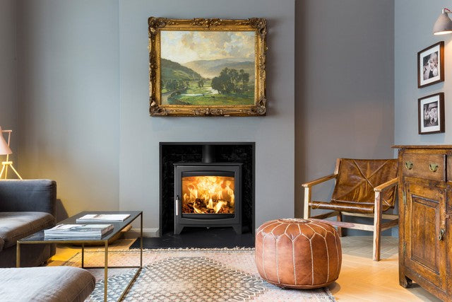 A black wood-burning fireplace with a glowing fire, set into a gray wall in a styled living room. A large framed landscape painting hangs above the fireplace, with a leather pouf, wooden chair, and cabinet nearby.
